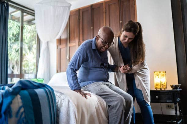 Une aide à domicile aidant un homme âgé à se lever de son lit dans sa chambre